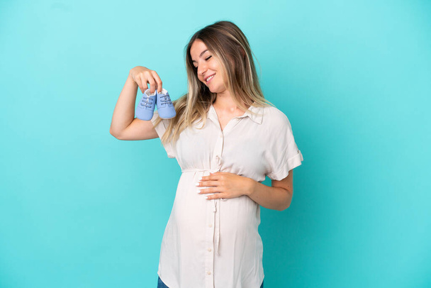 Νεαρή Ρουμάνα απομονωμένη σε μπλε φόντο έγκυος και κρατώντας μωρουδιακά μποτάκια - Φωτογραφία, εικόνα