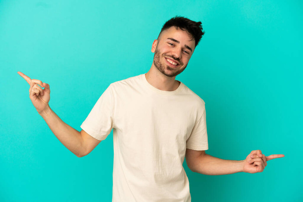 Junger kaukasischer Mann isoliert auf blauem Hintergrund, der mit dem Finger auf die Seitenlinien zeigt und glücklich ist - Foto, Bild