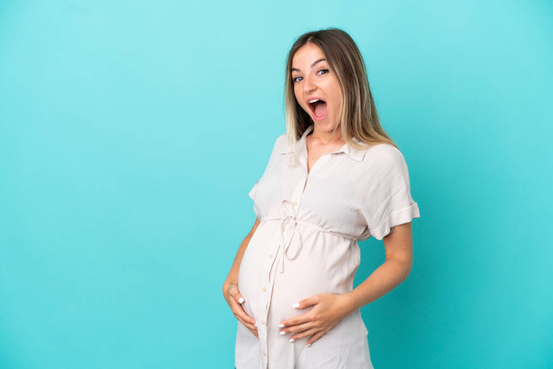 Young Romanian woman isolated on blue background pregnant and doing surprise gesture - Photo, Image