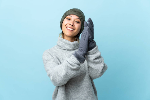 Młoda urugwajska dziewczyna w czapce zimowej odizolowana na niebieskim tle oklaskuje - Zdjęcie, obraz