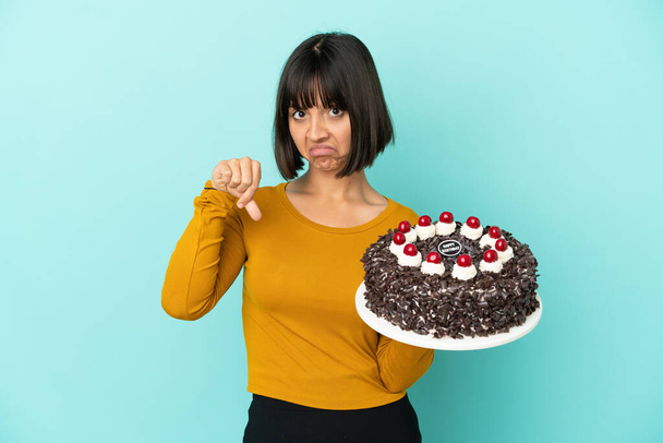 Giovane donna razza mista in possesso di torta di compleanno mostrando pollice verso il basso con espressione negativa - Foto, immagini