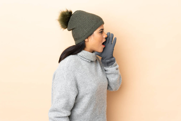 Молода колумбійська дівчина з зимовим капелюхом ізольована на бежевому фоні з відкритим ротом. - Фото, зображення