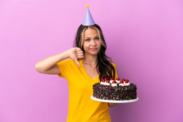 Adolescente rusa chica sosteniendo pastel de cumpleaños aislado en púrpura fondo mostrando el pulgar hacia abajo con expresión negativa - Foto, imagen