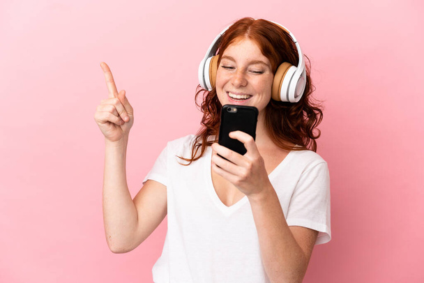 tinédzser vöröses nő elszigetelt rózsaszín háttér hallgat zenét egy mobil és énekel - Fotó, kép