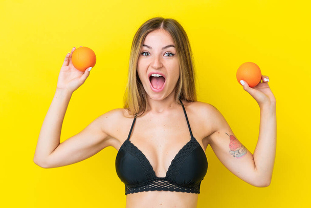 Young Romanian woman isolated on yellow background in swimsuit and holding an orange - Photo, Image