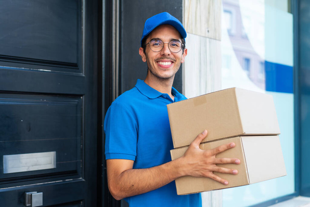 Молодой доставщик на открытом воздухе держит коробки с радостным выражением лица - Фото, изображение