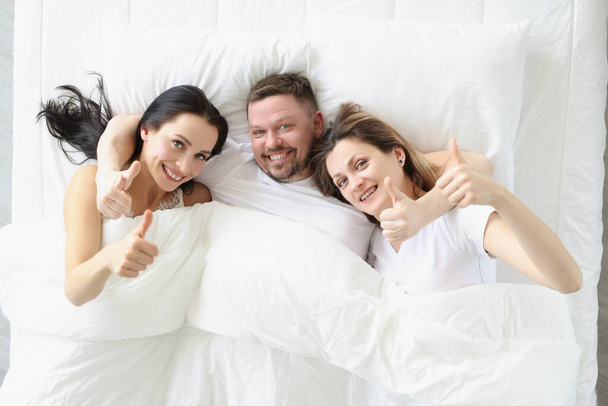 Two smiling women and man lie on bed and hold their thumbs up - Photo, Image