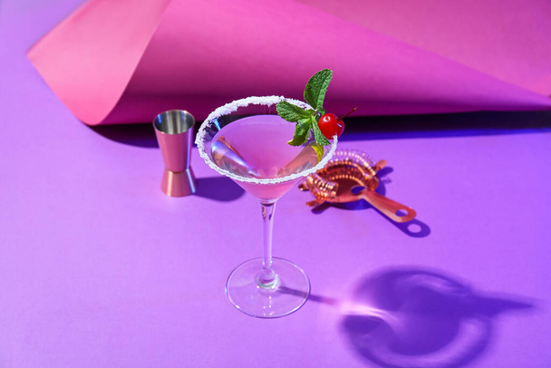 Glass of tasty cosmopolitan cocktail, shaker and strainer on color background - Fotó, kép