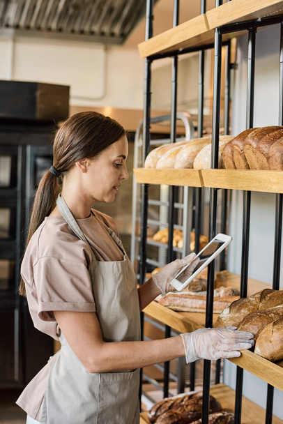 Young woman in apron standing by display with bread while using tablet - Photo, Image