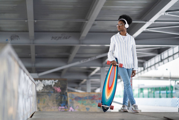 Trendy casual black woman in headphones hold longboard in skatepark or trendy urban space background - Photo, Image