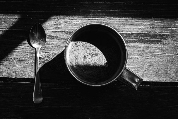 Zwart-wit foto, bovenaanzicht, grote metalen koffiemok en metalen lepel op een houten oppervlak met grijze kaneel, lichte schaduw, zonlicht 's morgens koffie - Foto, afbeelding