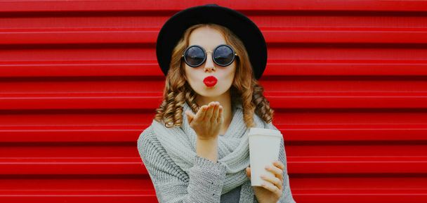 Portrait of beautiful young woman blowing her red lips sending air kiss with cup of coffee wearing a black round hat on red background - Foto, afbeelding