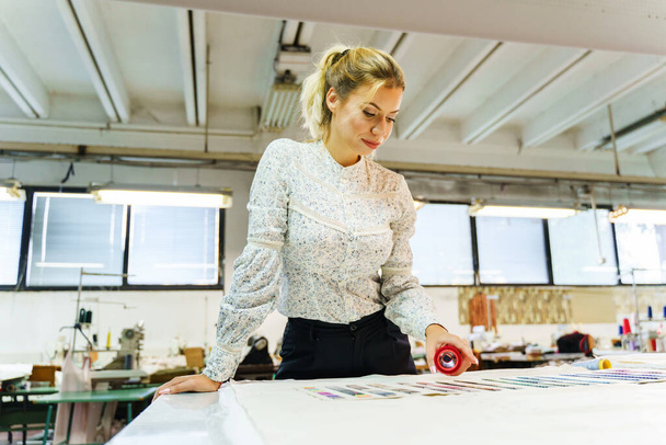 Junge erwachsene blonde Kaukasierin in der Schneiderei, die ein neues Designskizzenkleid auf Stoff schneidert - Unternehmerin arbeitet am Tisch - Konzept für die Gleichstellung der Geschlechter in kleinen Unternehmen - Foto, Bild