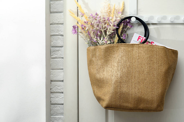 Stylish beach bag with beautiful bouquet of wildflowers and magazine hanging on white wooden door. Space for text - Фото, изображение
