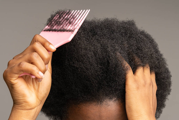 Close up of African female using comb, brushing curly trick hair isolated on studio gray wall.  - Photo, Image