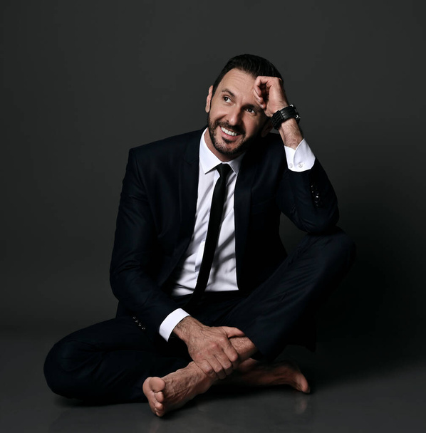 Smiling adult bearded man in black office suit sits barefooted on floor with legs crossed holding hand at head - Foto, immagini
