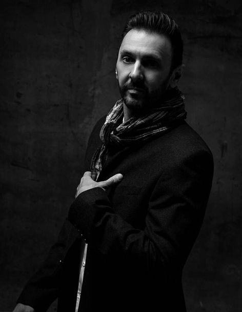 Black and white. Unshaved adult man in jacket and scarf stands sideways holding hand at chest and looks at camera - Fotoğraf, Görsel