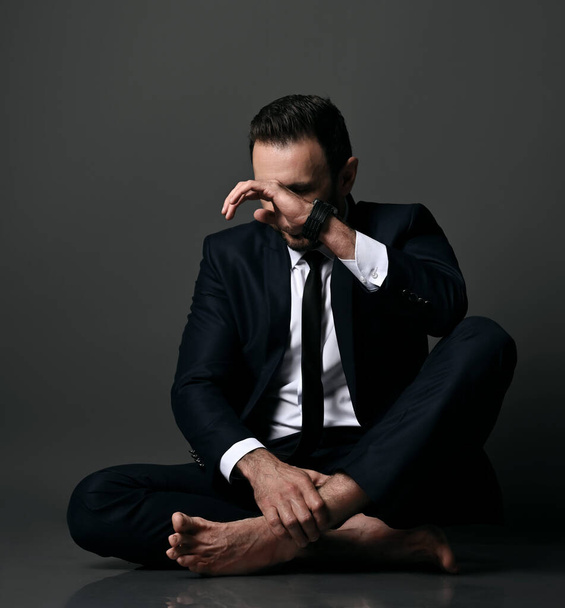 Failed adult man, bankrupt in black office suit sits barefooted on floor with legs crossed, closing face with hand - Foto, Bild