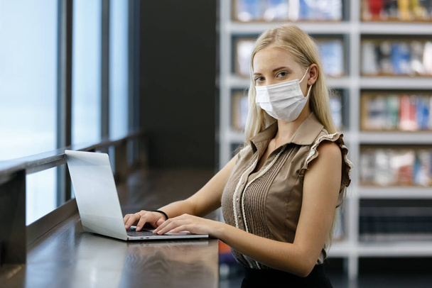 Belle femme aux cheveux blonds porter un masque médical à l'aide d'un ordinateur portable à la table de bureau. pigiste professionnel sur le lieu de travail. - Photo, image