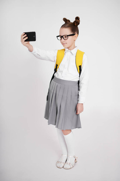 full length portrait of cute preteen girl in school uniform and eyeglasses taking selfie photo with smart phone over white background - Фото, изображение