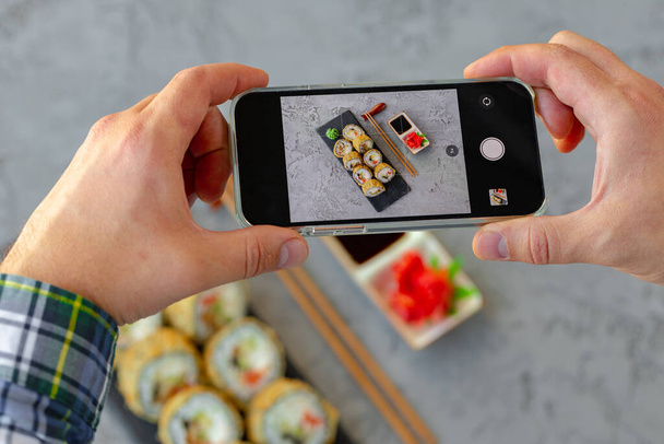 Male hands holding smartphone and taking a photo of sushi roll on gray background - Фото, изображение