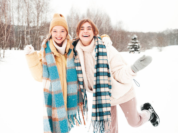Two young beautiful smiling hipster female in trendy warm clothes and scarfs.Carefree women posing in the street in park. Positive pure models having fun in snow. Enjoying winter moments. Xmas concept - Φωτογραφία, εικόνα