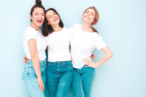 Three young beautiful smiling hipster female in trendy same summer white t-shirt and jeans clothes. Sexy carefree women posing near light blue wall in studio. Cheerful and positive models having fun - Fotoğraf, Görsel