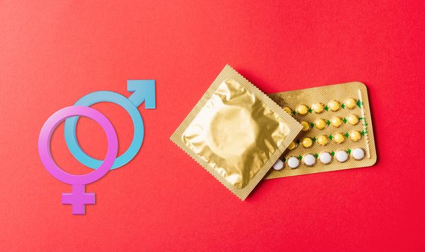World sexual health or Aids day. Male, female gender signs condom on wrapper pack and contraceptive pills blister hormonal birth control pills, studio shot isolated on a red background - Valokuva, kuva