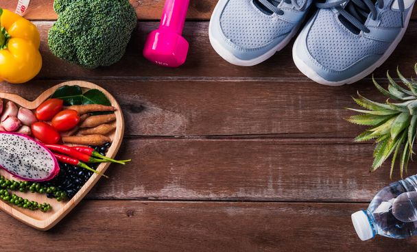 Top view of various fresh organic fruit and vegetable in heart plate and sports shoes, dumbbell and water, studio shot on wooden gym table, Healthy diet vegetarian food concept, World food day - 写真・画像
