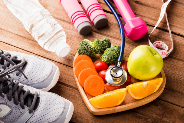 Top view of fresh fruits and vegetables in heart plate wood (apple, carrot, tomato, orange, broccoli) and sports equipment and doctor stethoscope on wooden table, Healthy lifestyle diet food concept - Photo, image
