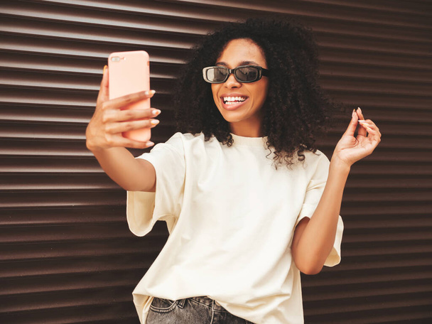 Beautiful black woman with afro curls hairstyle.Smiling hipster model in white t-shirt. Sexy carefree female posing in the street near brown wall in sunglasses.Looking at smartphone screen, using apps - Foto, imagen