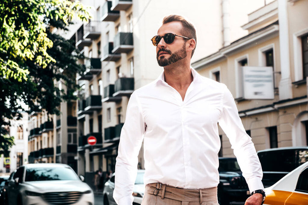 Portrait of handsome confident stylish hipster lambersexual model.Modern man dressed in white shirt and trousers. Fashion male posing in the street background in Europe city at sunset. In sunglasses - Zdjęcie, obraz