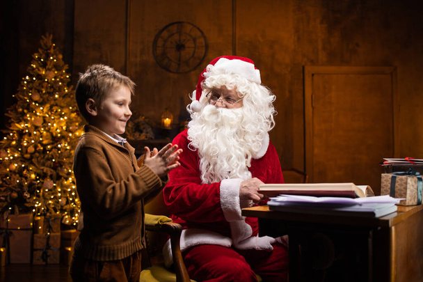 Santa Claus and little boy. Cheerful Santa is working while sitting at the table. Fireplace and Christmas Tree in the background. Traditional Christmas concept. - Fotografie, Obrázek