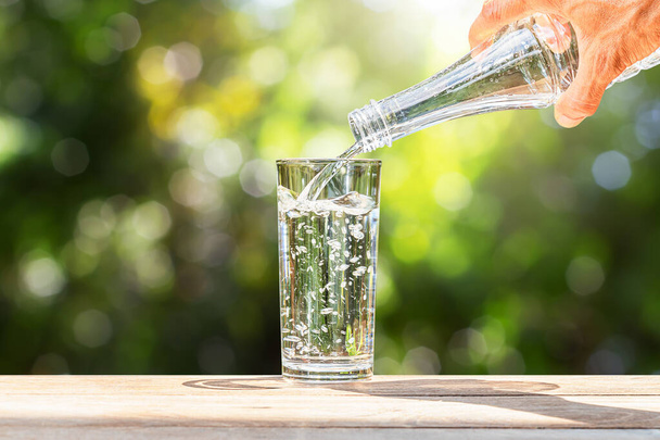 Ręczne trzymanie butelki wody pitnej wlewającej się do szkła na drewnianym stole na rozmazanym zielonym tle natury - Zdjęcie, obraz