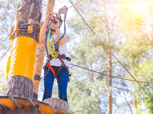 Kind mit orangefarbenem Helm klettert in Bäume auf Wald-Erlebnispark. Mädchen laufen auf Seilen und einer hohen Hängebrücke im sommerlichen Stadtpark. Extremsportausrüstung Helm und Karabiner - Foto, Bild