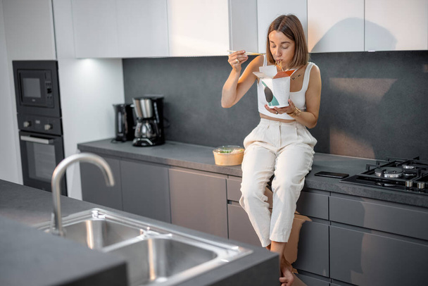 Γυναίκα τρώει takeaway τροφίμων, ενώ κάθεται στη σύγχρονη κουζίνα - Φωτογραφία, εικόνα