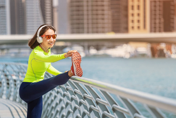 Szczęśliwa kobieta w słuchawkach rozciąga się i rozgrzewa po intensywnym treningu biegowym na nabrzeżu w Dubai Marina - Zdjęcie, obraz