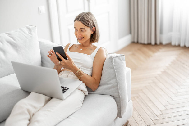 La mujer trabaja con el ordenador portátil y el teléfono en el sofá cómodo - Foto, Imagen