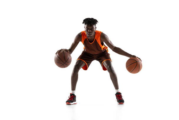 Joven africano, jugador de baloncesto en movimiento con dos bolas aisladas sobre fondo blanco del estudio. Vista frontal - Foto, Imagen