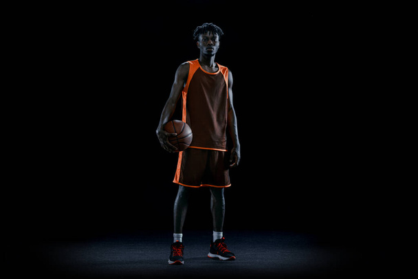 Jovem homem africano, jogador profissional de basquete posando com bola isolada sobre fundo escuro estúdio. - Foto, Imagem