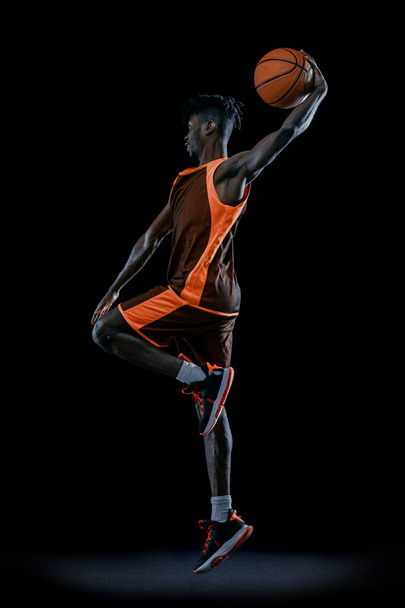 Vertikales Bild. Junger Afrikaner, Basketballspieler springt mit Ball isoliert über dunklen Studiohintergrund. - Foto, Bild