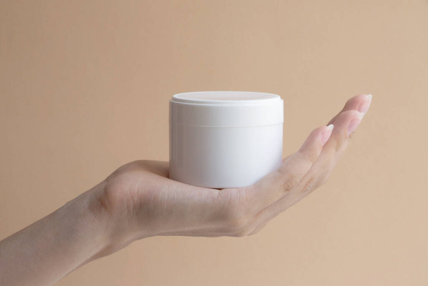 Mock-up of white cosmetic packaging is located on female palm. Close-up of round jar of body cream on beige background. Concept of beauty industry. - Φωτογραφία, εικόνα