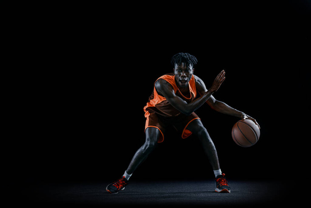 Z przodu. Młody Afrykanin, koszykarz ćwiczący z piłką na ciemnym tle studia. - Zdjęcie, obraz