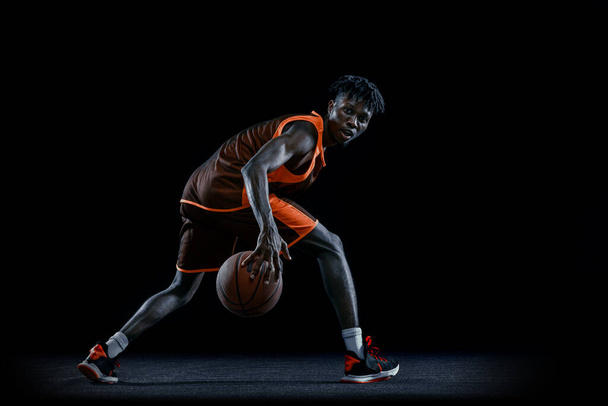 Jeden młody Afrykanin, koszykarz ćwiczący z piłką na ciemnym tle studia.. - Zdjęcie, obraz