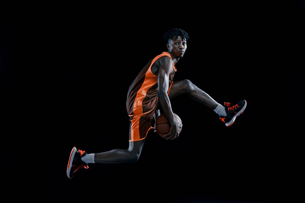 Widok z boku. Profesjonalny afrykański koszykarz ćwiczący z piłką odizolowaną na ciemnym tle studia. - Zdjęcie, obraz