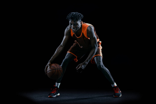 Portret młodego Afrykanina, koszykarza ćwiczącego z piłką na ciemnym tle studia. - Zdjęcie, obraz