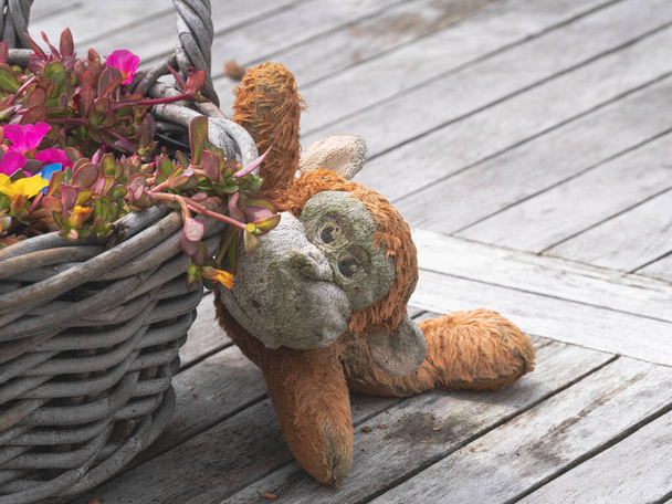 Vieux singe peluche altérée se trouve sur une table en bois à côté d'un panier de fleurs d'été - Photo, image