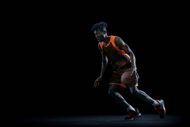 Profesjonalny afrykański koszykarz ćwiczący z piłką odizolowaną na ciemnym tle studia. - Zdjęcie, obraz