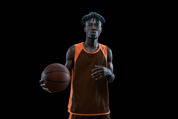 Prise de vue en studio d'un joueur de basket africain professionnel posant avec une balle isolée sur fond de studio sombre. - Photo, image