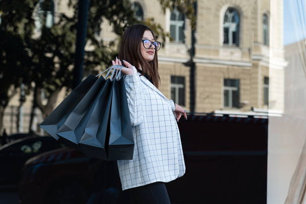 Молодая брюнетка с сумками для покупок. Девушка возле модного магазина после шопинга. Чёрная пятница. - Фото, изображение